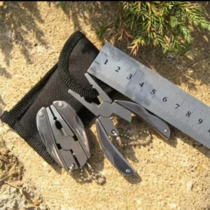 Folding keychain multi-tool with pliers beside a black pouch and a ruler on sand