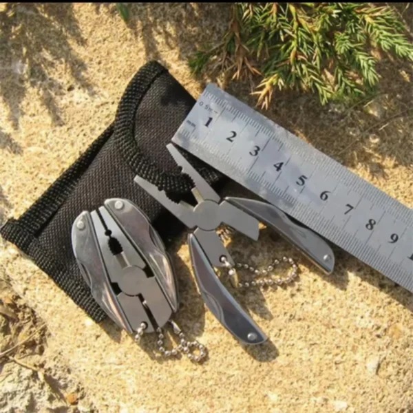 Folding keychain multi-tool with pliers beside a black pouch and a ruler on sand