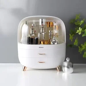White Ecoco cosmetic organiser with a clear domed lid, two drawers and rose-gold handles and feet, holding perfume and skincare bottles beside a green plant